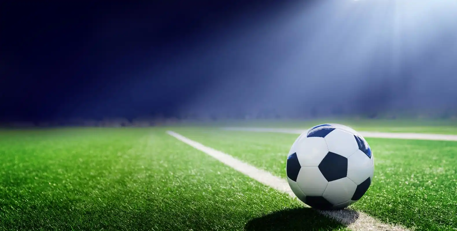 A soccer ball rests on a green field under bright stadium lights, casting a shadow across the white sideline.