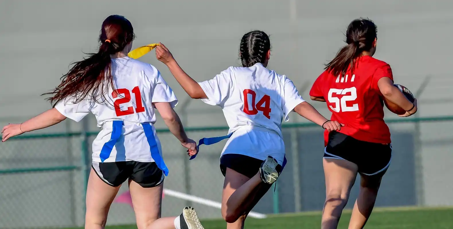 Three girls playing flag football, one in a red shirt holding the ball and two in white shirts, one grabbing a flag.