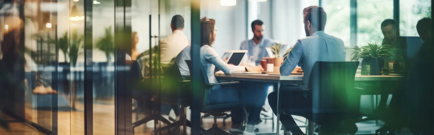 A group of people engaged in a meeting in a modern office space with large windows and green plants.