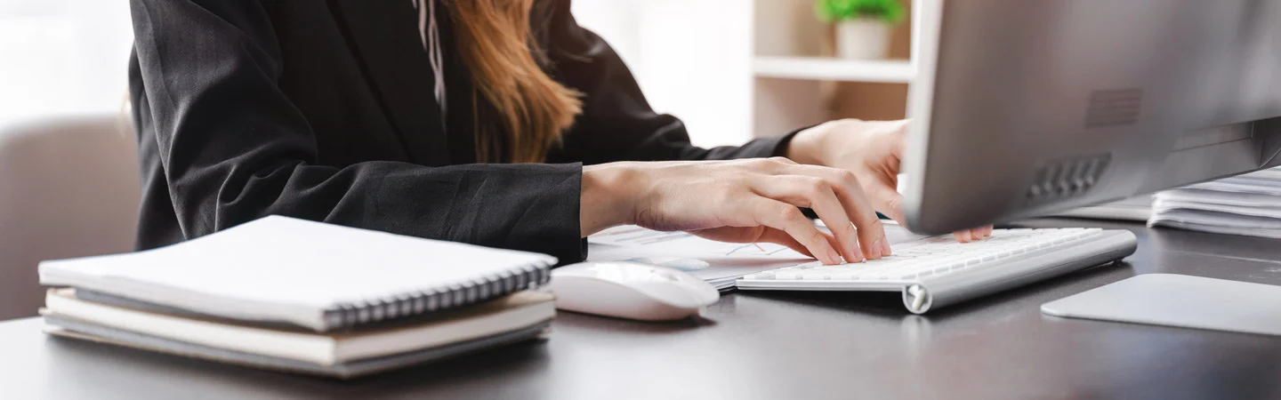 Person typing on a computer keyboard with notebooks and a mouse on a desk, taken from a side angle.