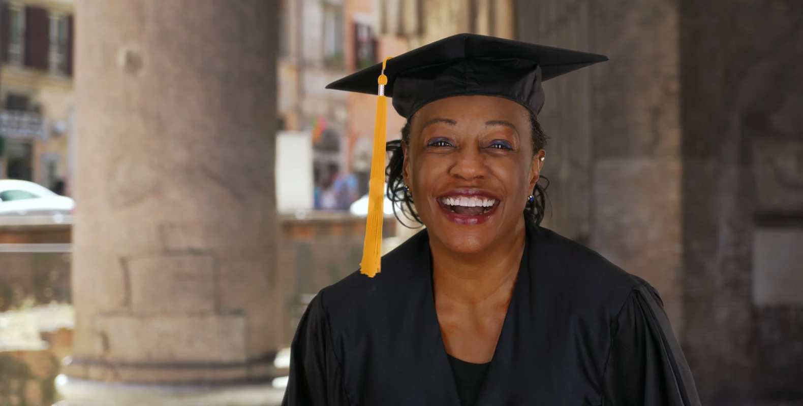 A smiling graduate, wearing a black cap and gown with a yellow tassel, stands in an outdoor setting.