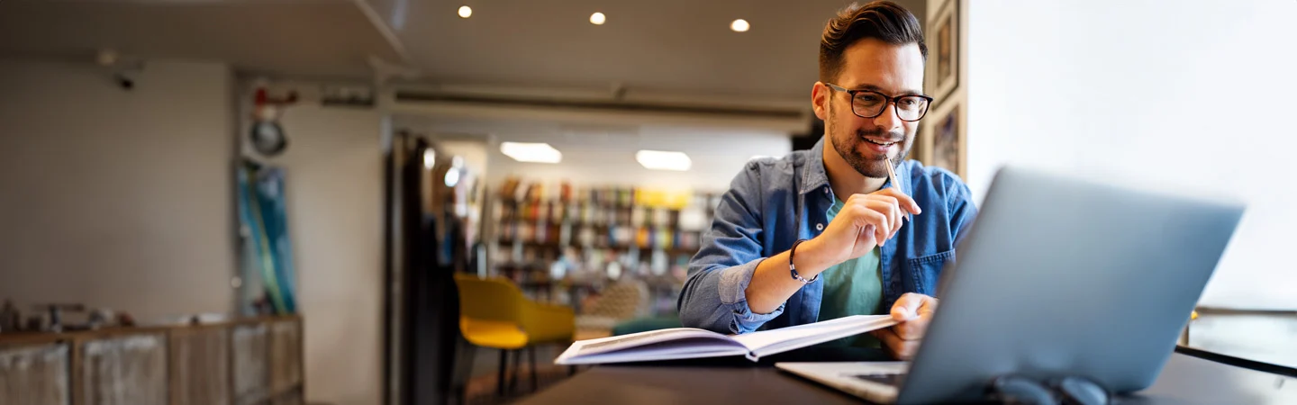 A man with glasses, sitting at a table in a library, working on a laptop and smiling while reading a book.
