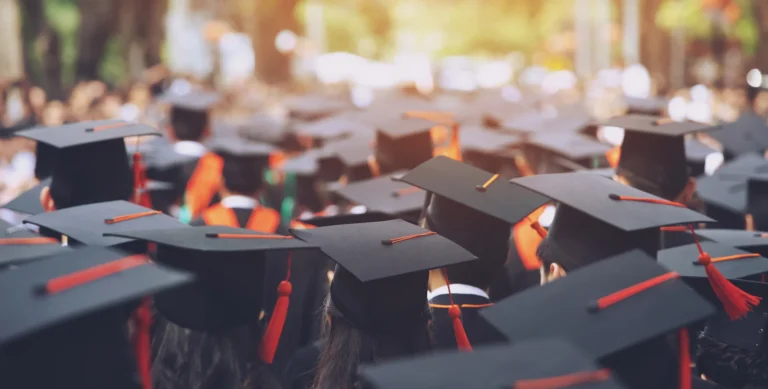 A large group of graduates in caps and gowns, seen from behind, during an outdoor graduation ceremony.