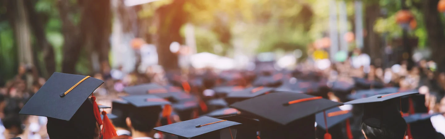 A crowd of graduates wearing black caps with tassels, gathered outdoors under bright sunlight.