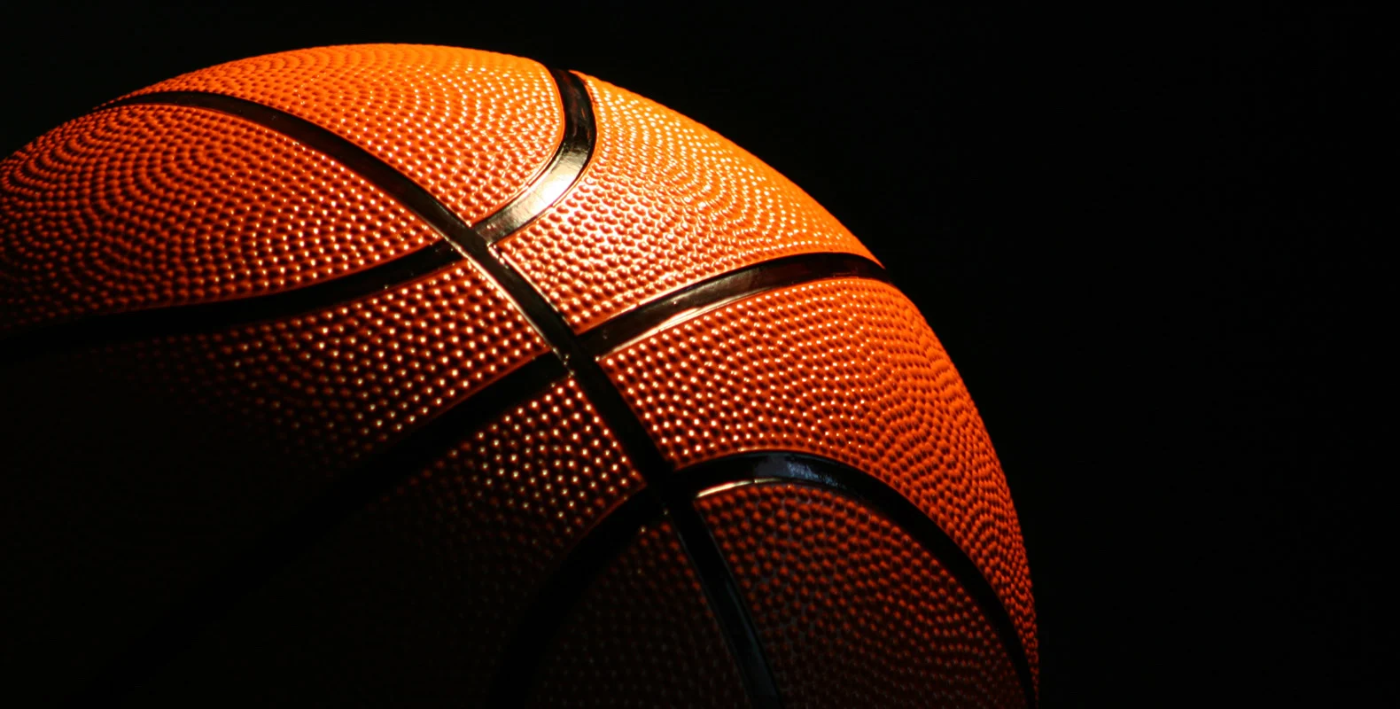Close-up of a basketball with dim lighting, highlighting its textured surface and lines against a black background.