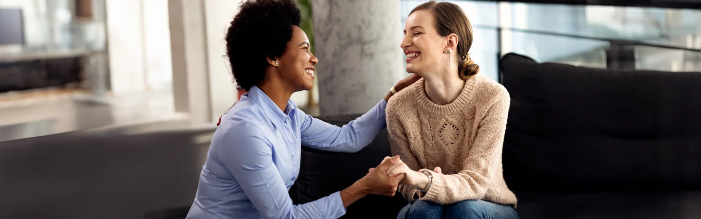 Two women smiling and holding hands while sitting on a couch, engaged in a friendly and supportive conversation.