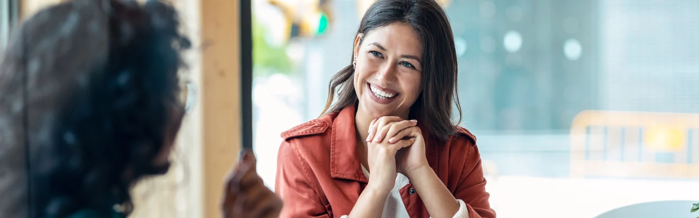 Person in a red jacket smiling and having a conversation with another person in a casual setting near a window.