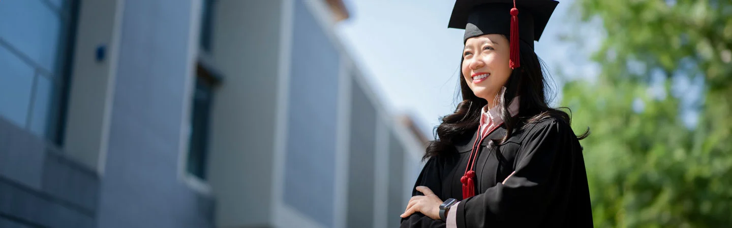 A graduate smiling outdoors, wearing a black cap and gown with red tassels.