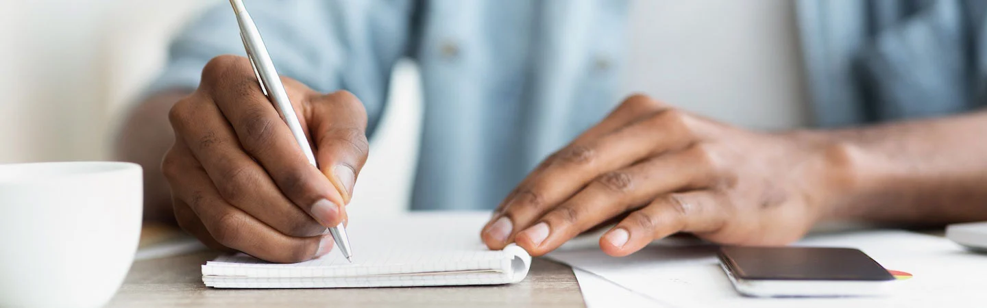 Close-up of a person writing in a notebook with a pen on a desk, with a smartphone and papers nearby.