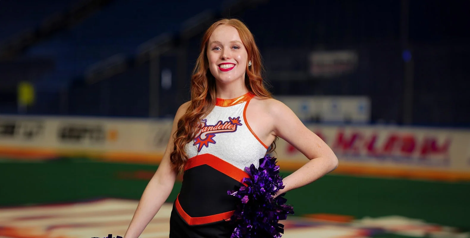 A cheerleader in a black, orange, and white uniform holds pom-poms, standing on a sports field with a bright smile.
