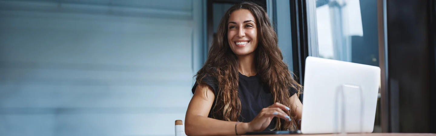 A smiling woman with long hair sits at a desk working on a laptop in a bright, modern setting.