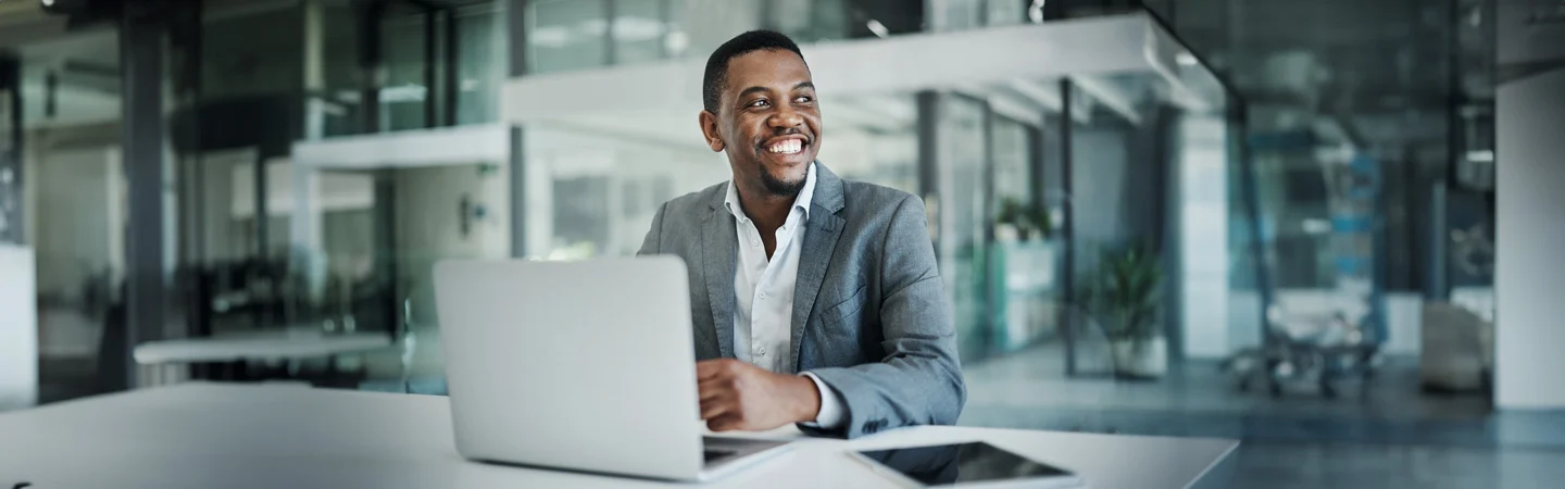 Man in a suit smiling while working on a laptop in a modern office with a tablet on the table.