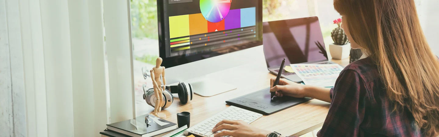 A person using a drawing tablet at a desk with a computer displaying a color wheel, surrounded by design tools.