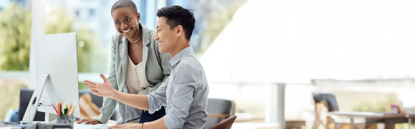 Two colleagues collaborate at a desk with a computer. The man is gesturing while the woman looks at the screen, smiling.