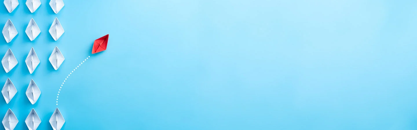 A red paper boat diverts from a group of white paper boats, symbolizing individuality and leadership on a light blue background.