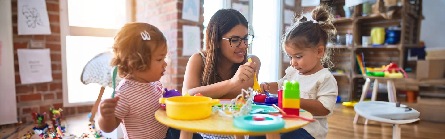 A woman plays with two young girls at a table filled with colorful toys in a bright room.