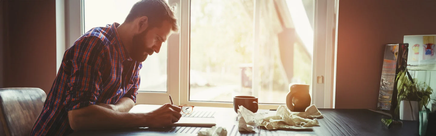 A bearded man writes in a notebook at a desk, with a window and coffee cup nearby. The room is softly lit by natural light.