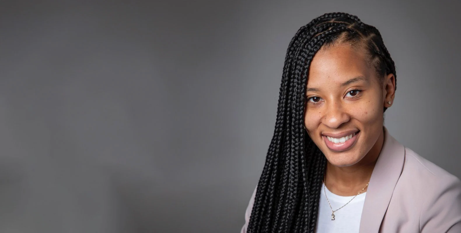 Woman with long braided hair wearing a light blazer and smiling against a gray background.