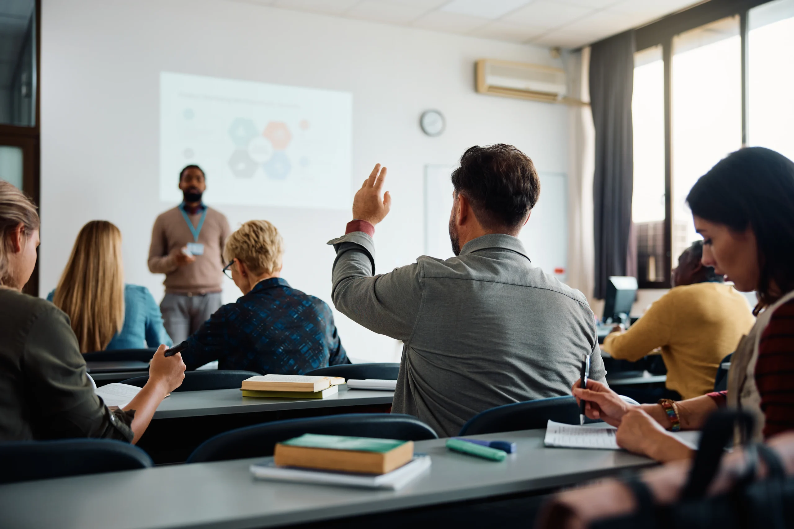 A man raises his hand in a classroom while others listen to a lecturer in front of a projector screen.