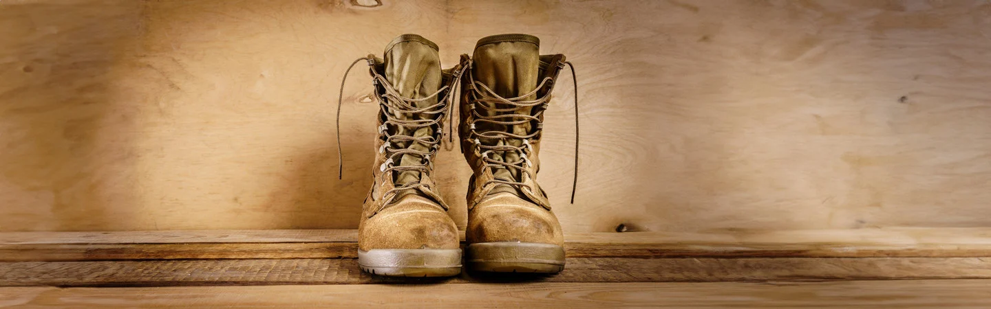 A pair of worn tan combat boots with laces, placed against a wooden backdrop.