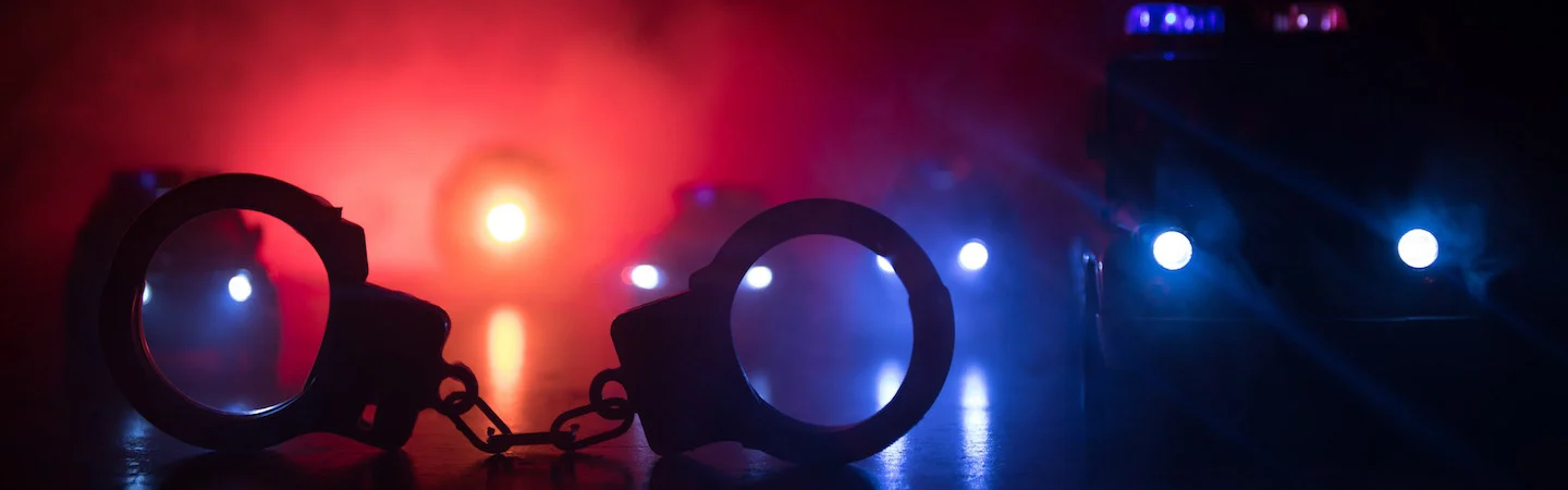 Handcuffs on the ground with police car lights flashing in the background in a dark, foggy scene.