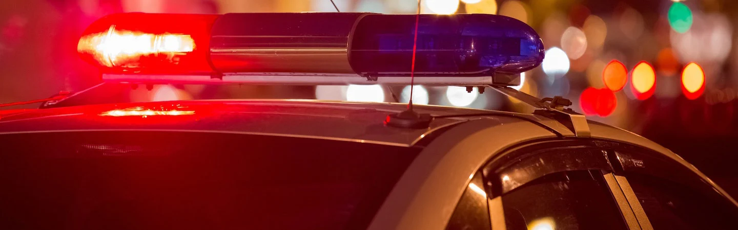 Police car with red and blue lights flashing at night, blurred city lights in the background.
