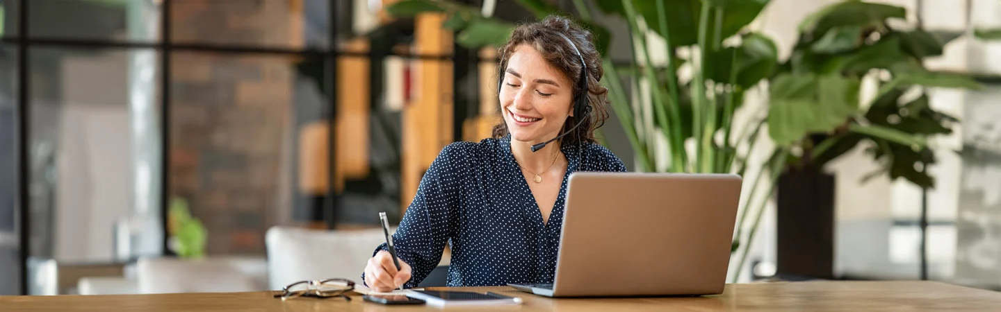 A smiling woman wearing a headset works on a laptop in a bright, modern office space with plants in the background.