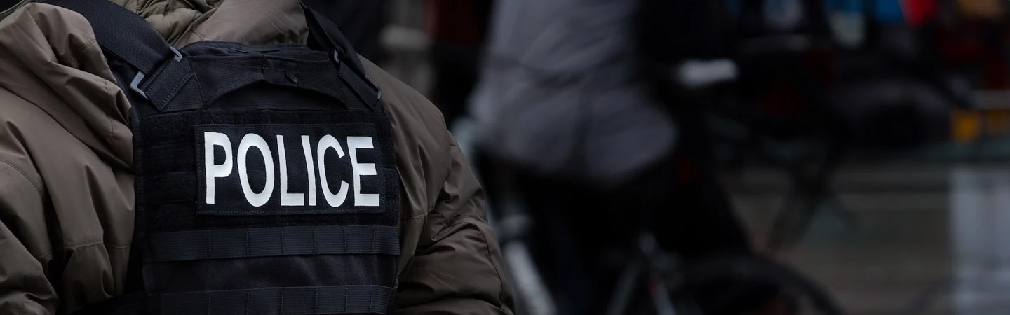A person dressed in a jacket with a vest labeled "POLICE" stands facing away, with blurred figures in the background.