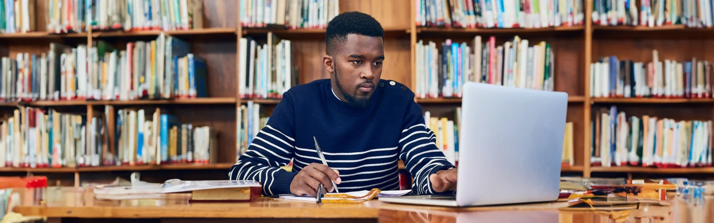 A man sitting at a library table, writing in a notebook while looking at his laptop. Bookshelves are in the background.