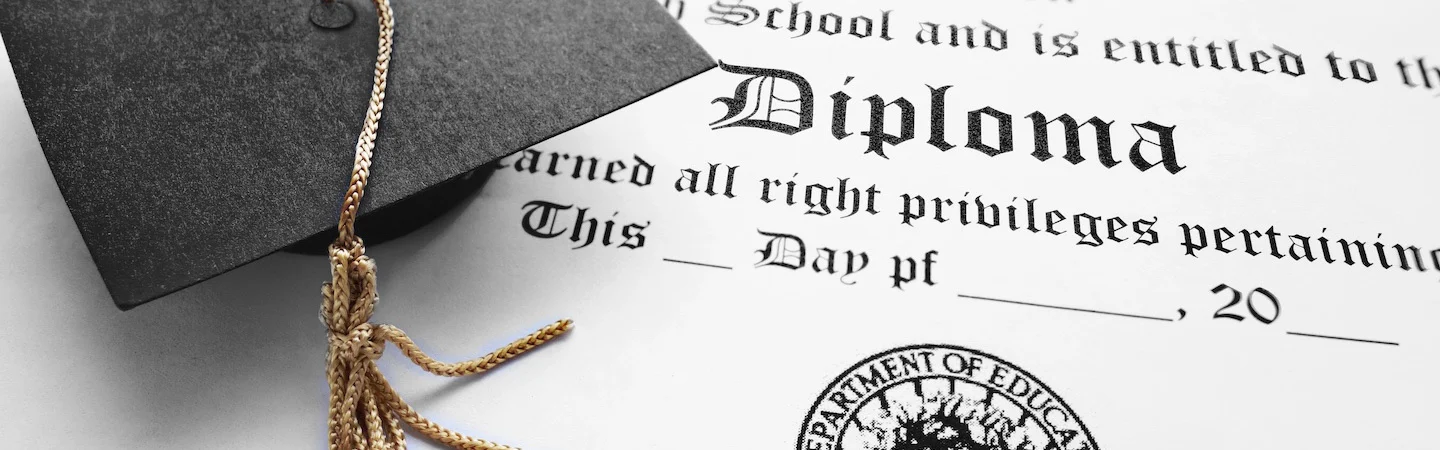 A close-up of a diploma next to a black graduation cap with a gold tassel.