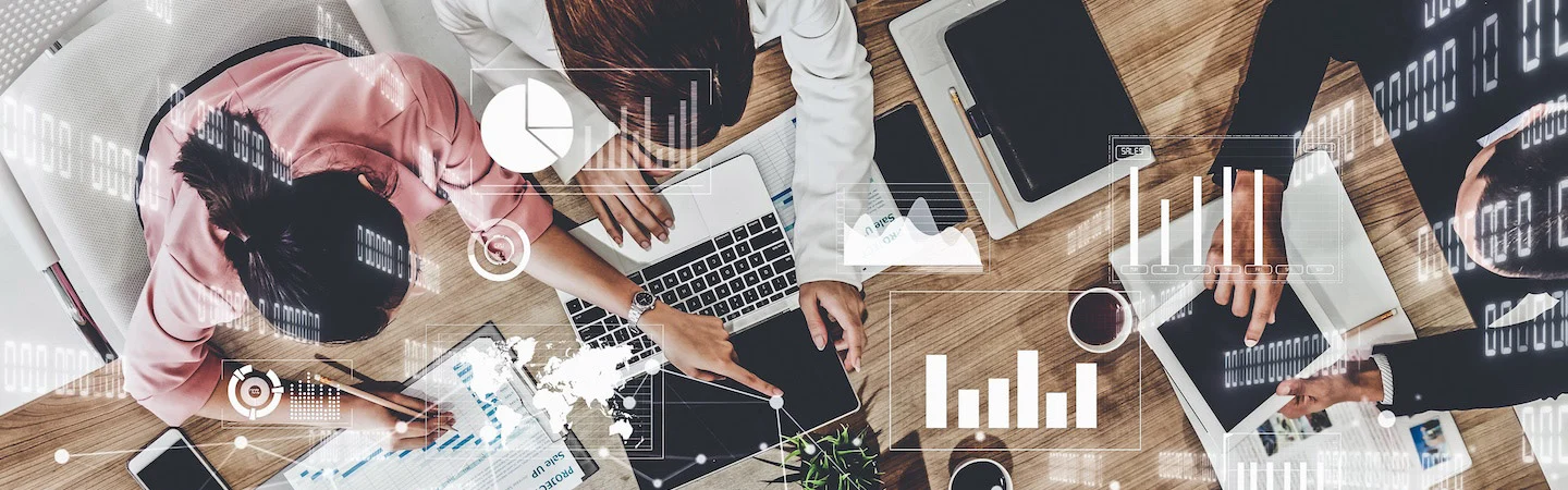 Aerial view of four people working on laptops and tablets at a desk, surrounded by data charts and graphs.