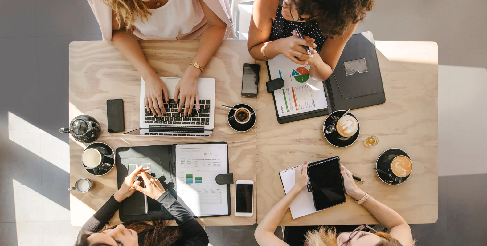 Four people sit around a table with laptops, tablets, coffee, and documents, engaging in a collaborative work session.