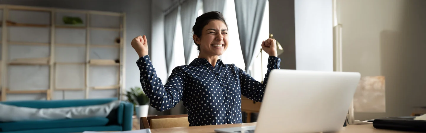 Person at a desk looking at a laptop, smiling and raising their fists in a celebratory gesture.