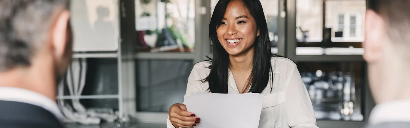 A woman holding papers and smiling while sitting in front of two people during a meeting in an office setting.