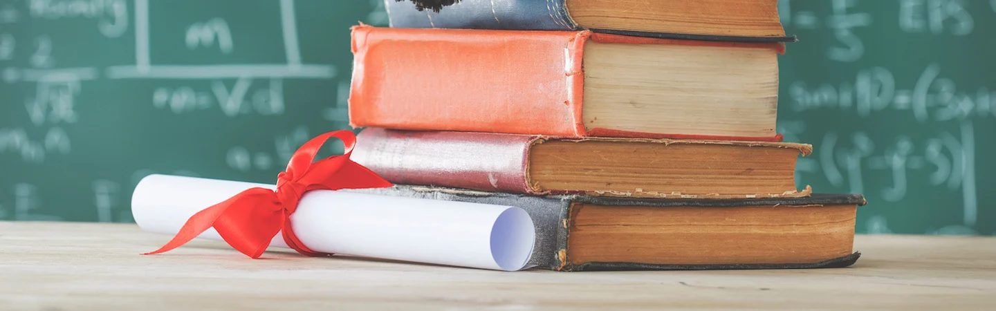 Stack of books beside a rolled diploma tied with a red ribbon on a table, with a chalkboard in the background.