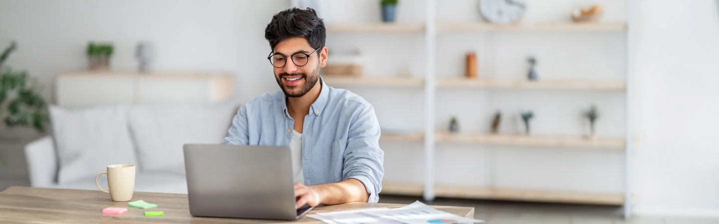 A man wearing glasses and smiling while working on a laptop at a desk with a mug and documents in a well-lit room.