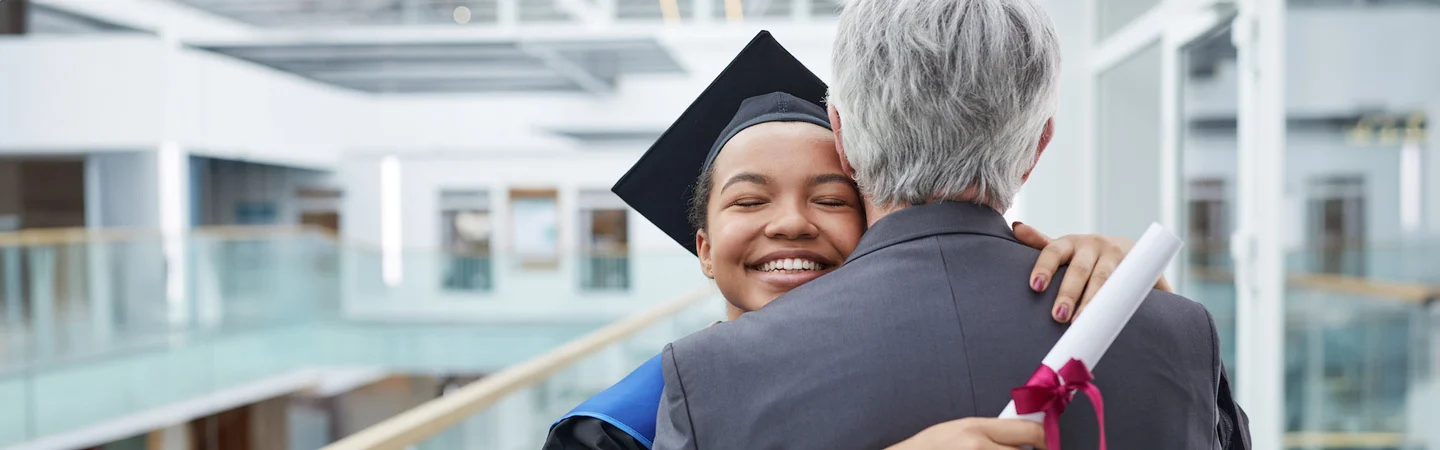 Graduate in cap and gown hugging an older person while holding a diploma with a red ribbon in a modern building.