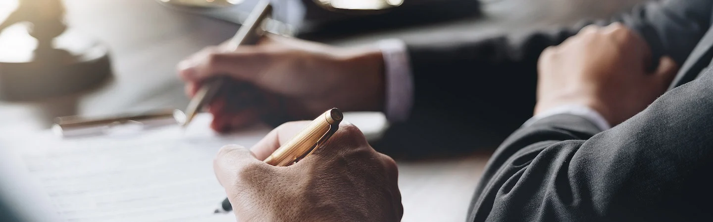 Close-up of hands in suits writing on documents at a desk, suggesting a professional or business setting.