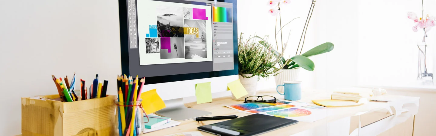 A cluttered desk with a computer displaying graphic design software, various stationery items, and a potted plant.