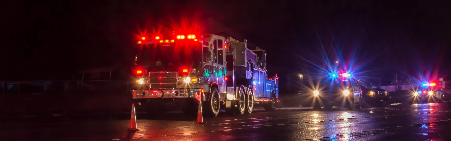 Night scene with a fire truck and police cars with flashing lights parked on a wet road. Traffic cones are set up.