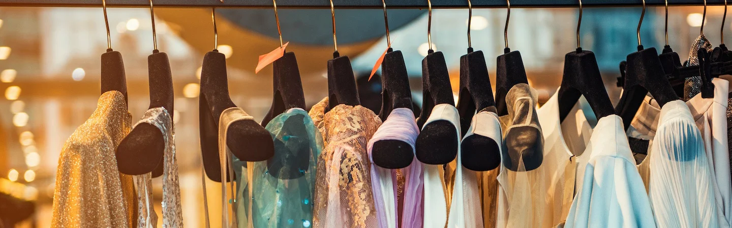 A row of assorted dresses hanging on black hangers in a clothing store, with blurred lights in the background.