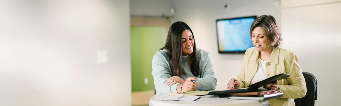 Two women sitting at a table, reviewing documents together in a bright office setting.
