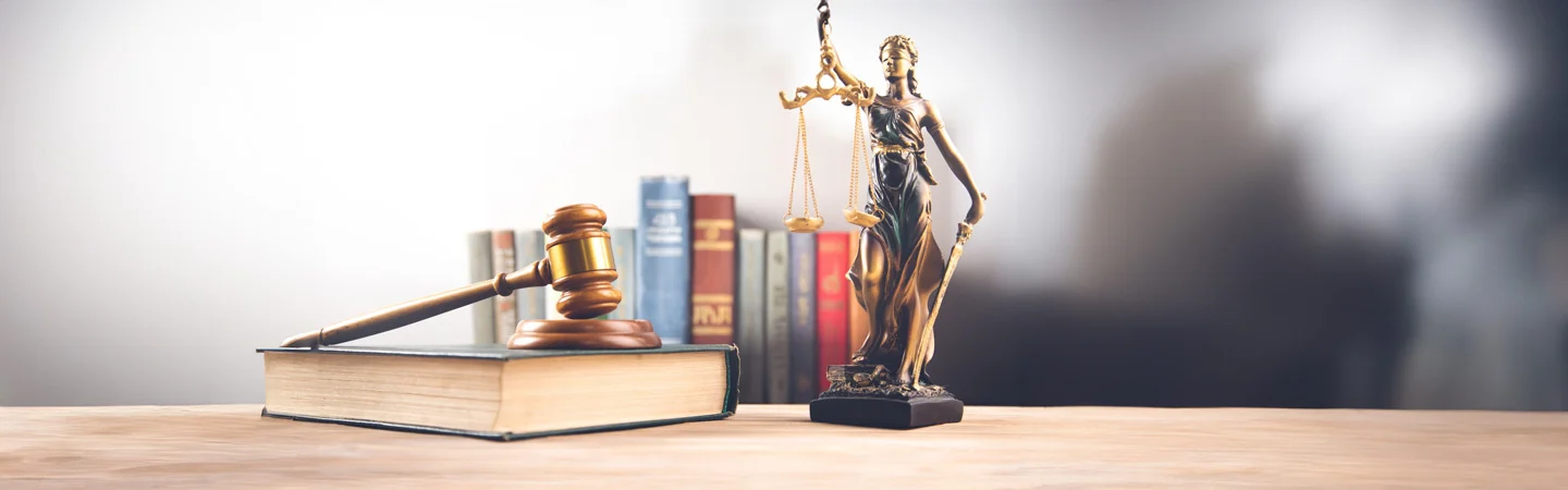 A judge's gavel rests on a book beside a Lady Justice statue with a scales of justice in front of several books.