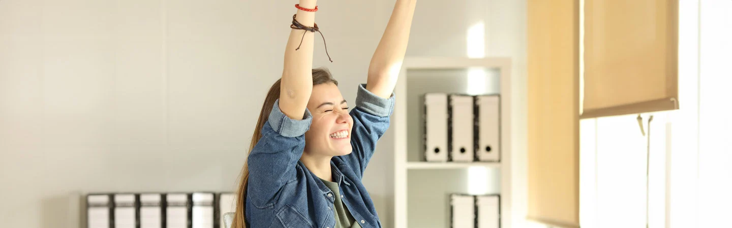 A person in a denim shirt smiles and raises their arms in celebration, standing in a bright office space.