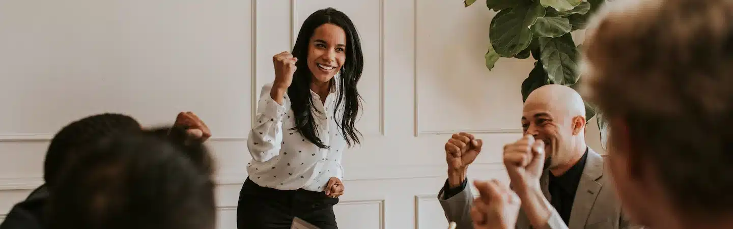A group of people celebrates in a meeting room. A woman stands in the center with a raised fist, smiling.