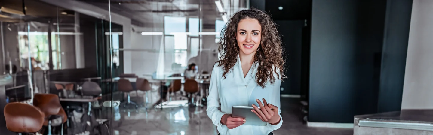 A woman with curly hair, wearing a white blouse, smiles while holding a tablet in a modern office space.