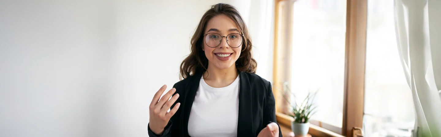 Smiling person with glasses and wavy hair, wearing a black jacket and white shirt, standing by a window with plants.