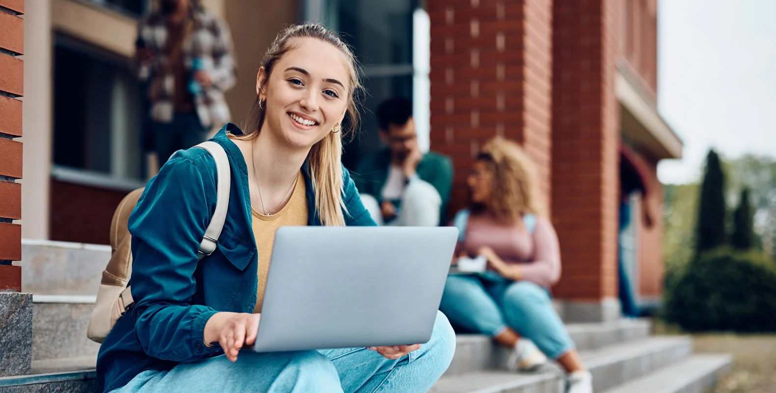 Smiling student sitting on outdoor steps with a laptop, other students in the background near a brick building.