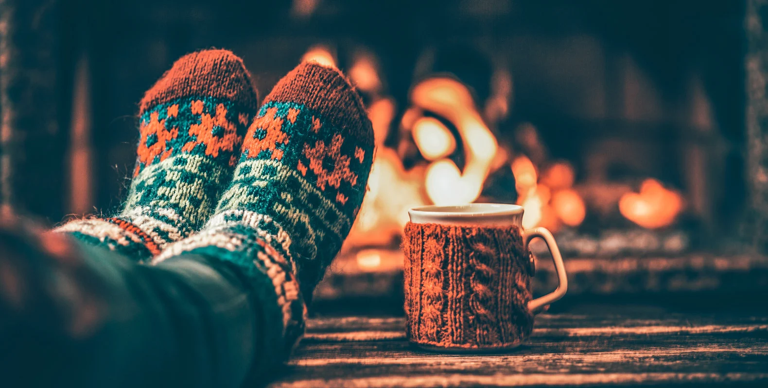 Cozy scene with person wearing knitted socks, relaxing by a fireplace, and a cup of hot beverage in a knitted cozy beside them.