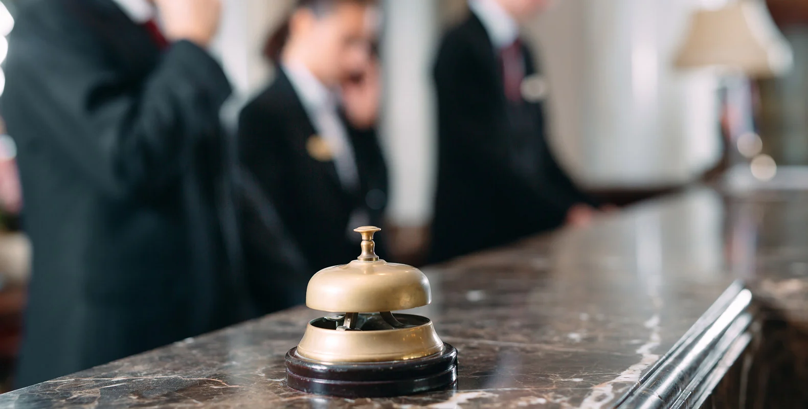 Close-up of a brass service bell on a hotel check-in counter, with three blurred staff members in the background.